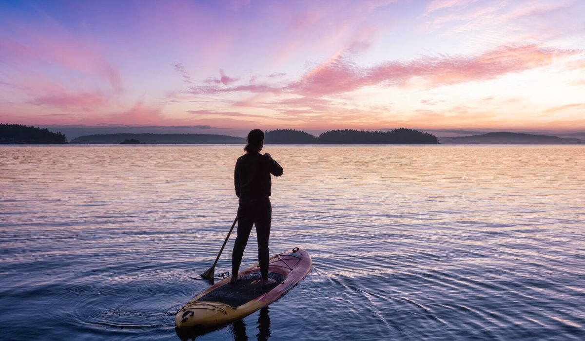 Adventurous Caucasian Adult Woman on a Stand Up Paddle Board is paddling on the West Coast of Pacific Ocean. Sunny Sunrise Sky Art Render. Victoria, Vancouver Island, BC, Canada.